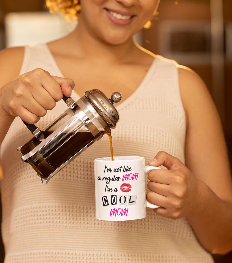 A stylish mom wearing cozy loungewear holds the "I'm Not Like a Regular Mom, I'm a Cool Mom" mug while pouring coffee. Her joyful expression captures the fun and lighthearted spirit of this coffee cup, making it a perfect gift for cool moms everywhere.