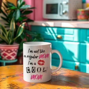A high-quality white ceramic coffee mug with bold black script that reads "I'm Not Like a Regular Mom, I'm a Cool Mom," sitting on a cozy wooden table with a steaming cup of coffee, making it the perfect funny and thoughtful gift for moms who love caffeine.