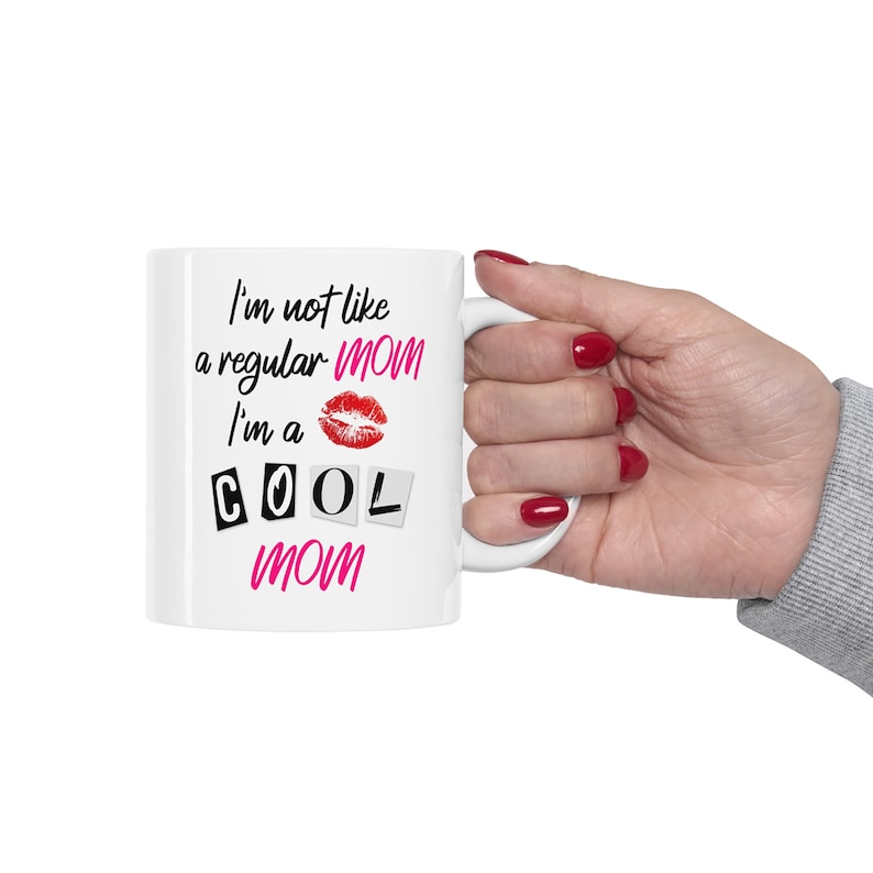 A white ceramic coffee mug that has the words from Mean Girls, Im not like a regular mom, Im a cool mom, printed on it. The glossy cup is being held up by a woman with red nail polish.