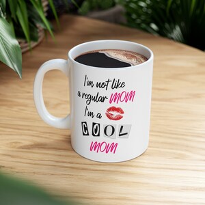 A white ceramic mug that appears to be of high quality is set atop a white oak wooden surface, with greenery surrounding it. The cup contains hot coffee, and has the mean girls quote, I'm not like a regular mom, I'm a cool mom written on it.