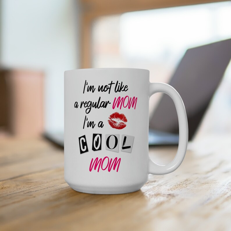15 ounce white ceramic coffee mug with the mean girls quote: I'm not like a regular mom, I'm a cool mom, set atop a wooden desk with a laptop in the background.