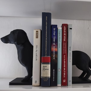 Puede incluir: Una estantería con sujetalibros con forma de perro negro que sostienen una colección de libros. Los libros tienen lomos y títulos de varios colores. Un jarrón blanco con una parte superior decorativa está en el lado derecho.
