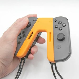 Könnte beinhalten: Ein Paar orangefarbene und graue Nintendo Switch Joy-Con-Controller, die an einem schwarzen Band befestigt sind. Die Controller befinden sich in einer Handheld-Konfiguration, wobei die linken und rechten Joy-Cons miteinander verbunden sind.