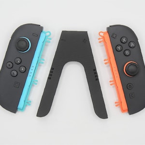 Op de afbeelding: Een zwarte gamecontroller met blauwe en oranje accenten, en een zwarte standaard. De controller heeft knoppen en een joystick. De standaard heeft de vorm van een omgekeerde "V".
