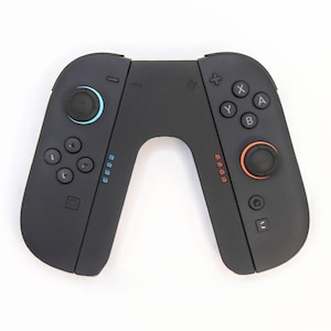 May include: A black games controller with blue and orange detailing. The controller is split into two sections, joined by a curved section. Each part has a joystick, buttons, and indicator lights. The buttons are labelled with letters and symbols.