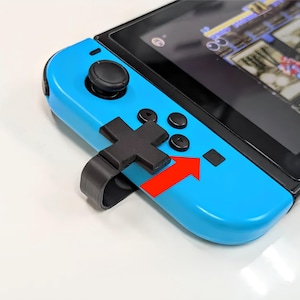 Pode incluir: Um console Nintendo Switch azul com um acessório de controlador preto. O acessório tem um botão em forma de cruz e um botão quadrado. O console está em uma superfície branca.