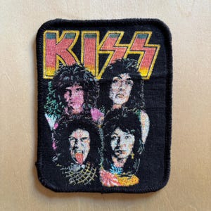 Pode incluir: Um emblema retangular de tecido preto com uma estampa gráfica da banda de rock KISS. A palavra KISS é exibida em letras grandes, estilizadas e amarelas e vermelhas no topo, acima de um retrato dos quatro membros da banda com cabelos escuros.