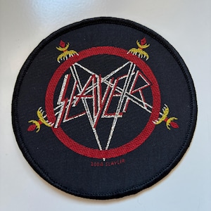 May include: A round, black patch featuring the word "SLAYER" in a white, angular font, encircled by a red ring. The design includes yellow flame-like accents and the text "2004 SLAYER" at the bottom. The patch has a stitched border.