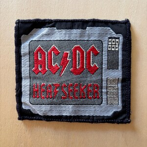 May include: A square embroidered patch featuring a black border and a gray center panel. The text AC/DC and HEAT SEEKER are stitched in red with a lightning bolt symbol. The design includes a gray grid pattern and two rectangular gray sections.