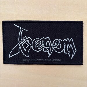 Op de afbeelding: Een rechthoekige zwarte geborduurde patch met de witte gestileerde tekst Venom in een grillig, scherp lettertype. Onder het hoofdlogo staat in kleinere witte letters 1996 VENOM, UNDER LICENSE TO RAZAMATAZ. De patch heeft een dikke zwarte rand.