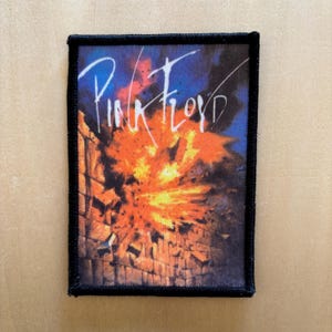 May include: A rectangular fabric patch with a black stitched border featuring a graphic of a brick wall exploding into orange and yellow flames against a dark blue sky. The text Pink Floyd is written in white cursive script across the top.