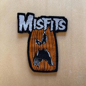 Könnte beinhalten: Ein gestickter Aufnäher mit dem Wort MISFITS in weißen Blockbuchstaben über einem orangefarbenen Kürbis mit einem schwarzen geschnitzten Gesicht. Das Design hat einen schwarzen Rand und detaillierte vertikale Linien auf dem Kürbis.