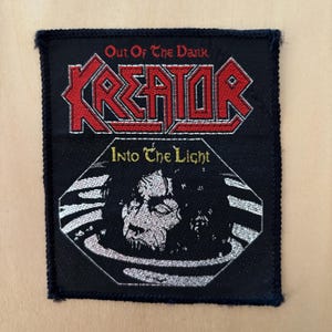 Op de afbeelding: Een rechthoekige zwarte geborduurde patch met de rode tekst Kreator boven de witte tekst Out Of The Dark. Daaronder staat de tekst Into The Light boven een witte afbeelding van een gezicht dat uit een cirkelvormig kader op een zwarte achtergrond komt.