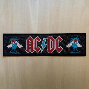 Könnte beinhalten: Gestickter schwarzer Aufnäher mit dem Bandnamen "AC/DC" in Rot und einem blauen Blitz. Der Aufnäher hat zwei cartoonartige, insektenartige Figuren auf beiden Seiten mit blauen Körpern, weißen Flügeln und roten Augen.