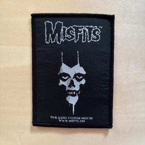 Può includere: Una toppa rettangolare in tessuto nero con il logo bianco Misfits in alto e una grafica bianca di un teschio al centro. Il testo in basso recita TM & © 2002 CYCLOPIAN MUSIC INC WWW.MISFITS.COM. La toppa presenta un bordo nero cucito.