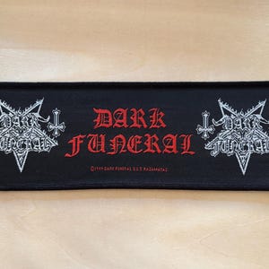 Può includere: Toppa rettangolare nera con le parole "DARK FUNERAL" in lettere gotiche rosse. La toppa presenta disegni di pentagramma e croce bianchi su entrambi i lati del testo. Il testo "©1999 DARK FUNERAL U.S.T. RAZAMATAZ" è in basso.