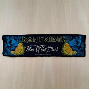 Puede incluir: Un parche rectangular negro bordado con el texto amarillo IRON MAIDEN y el texto blanco Fear Of The Dark. El diseño incluye ilustraciones gráficas azules y amarillas de un rostro a ambos lados y el texto 2004 IRON MAIDEN HOLDINGS LTD.