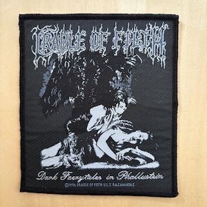 May include: A black and white patch with the band name "Cradle of Filth" in a gothic style. The patch features an illustration of figures and the text "Dark Faerytales in Phallustein".