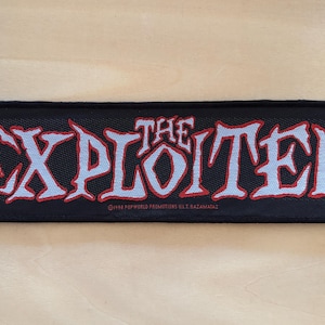 Può includere: Toppa rettangolare nera con la scritta "THE EXPLOITED" in un carattere stilizzato azzurro chiaro con contorno rosso. La toppa include anche il testo in basso: ©1998 POPWORLD PROMOTIONS U.K. RAZAMATAS.
