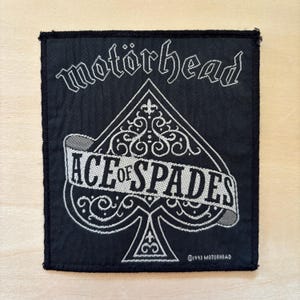 以下が含まれることがあります： 白い刺繍が施された長方形の黒い布製ワッペン。上部にはスタイリッシュなフォントで「motörhead」の文字。その下には大きなスペードのシンボルがあり、その中に「ACE of SPADES」と「©1993 MOTORHEAD」と書かれたバナーが配置されています。