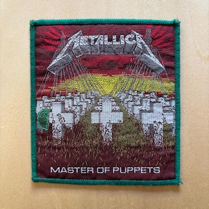 以下が含まれることがあります： 上部にMetallicaのロゴ、下部にMASTER OF PUPPETSのテキストが織り込まれた長方形のワッペン。赤と黄色の空の下に白い十字架が並ぶ墓地が描かれており、周囲は緑色のステッチで縁取られている。