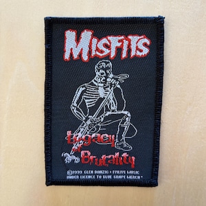 以下が含まれることがあります： 黒い長方形の布製ワッペン。白い刺繍でマイクを持って膝をつく骸骨が描かれている。上部には赤と白でMISFITSの文字、その下には赤でLegacy Brutalityの文字があり、下部には著作権に関するテキストが記載されている。