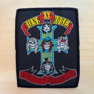 May include: Black rectangular patch featuring the Guns N' Roses logo with a cross design. The cross is adorned with skull illustrations and the words "GUNS N' ROSES" and "APPETITE FOR DESTRUCTION" in red and yellow.
