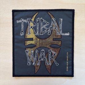 May include: Square patch featuring the text "TRIBAL WAR" in a bone-like font, set against a gold and brown design. The patch has a dark blue background and a black border. The text "©2003 BLUE GRAPE LTD" is at the bottom right.