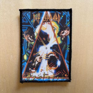 May include: A rectangular fabric patch featuring the Def Leppard Hysteria album cover art. The design includes a large orange triangle, blue geometric patterns, and portraits of band members against a white background with black stitched edges.