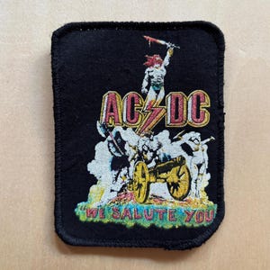 May include: A rectangular black embroidered patch featuring the AC/DC logo in red and yellow. The design includes a central figure holding a sword, a cannon on wheels, and the text "WE SALUTE YOU" in red and yellow lettering at the bottom.