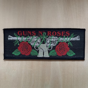 Può includere: Una toppa rettangolare nera ricamata con la scritta GUNS N' ROSES in rosso. Il design include due revolver argentati intrecciati con viti spinose verdi e due grandi rose rosse con foglie verdi posizionate sotto le armi.