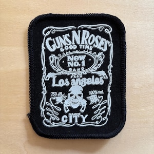May include: A rectangular black fabric patch with white embroidered text and graphics. The design features the text GUNS N ROSES, GOOD TIME, NEW NO.1 BAND, FROM LOS ANGELES, 750 dB, 100% vol., and PARADISE CITY, alongside a skull and two roses.