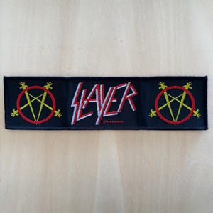 Op de afbeelding: Een rechthoekige zwarte geborduurde patch met het woord Slayer in witte en rode letters. Aan zowel de linker- als rechterkant staat een geel pentagramsymbool in een rode cirkel. De tekst 2004 SLAYER is onderaan duidelijk zichtbaar.