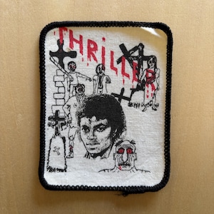 May include: A rectangular fabric patch with a black stitched border on a white background. It features a black and white portrait of a person, surrounded by illustrations of figures, crosses, and a tombstone. The red text THRILLER is printed above.