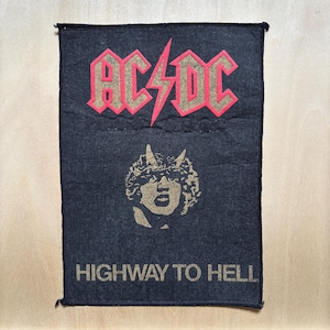 Puede incluir: Parche rectangular negro con el nombre de la banda "AC/DC" en letras rojas y doradas con un rayo. Debajo, una imagen dorada de una figura con cuernos y el texto "HIGHWAY TO HELL" en dorado.