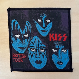 Könnte beinhalten: Ein quadratischer Stoffaufnäher mit einer Grafik der Band KISS mit blauen Gesichtern und schwarzem Make-up. Der Text KISS erscheint in Rot und die Worte BRITISH TOUR sind unten links in Rot aufgedruckt. Der Aufnäher hat einen schwarzen Rand.