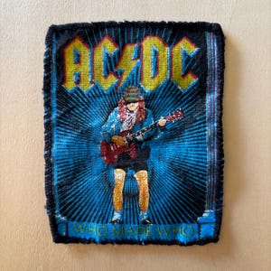 May include: A rectangular embroidered patch featuring the AC/DC logo in yellow at the top, with a central figure playing a red electric guitar. The background is a blue sunburst pattern, and the text WHO MADE WHO is printed in yellow at the bottom.