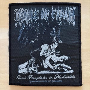 Könnte beinhalten: Schwarzer quadratischer Aufnäher mit einer detaillierten Illustration in Grau- und Weißtönen. Der Text "CRADLE OF FILTH" steht oben, mit den Worten "Dark Faerietales in Phallustein" und Copyright-Informationen unten. Der Aufnäher hat einen schwarzen Rand.
