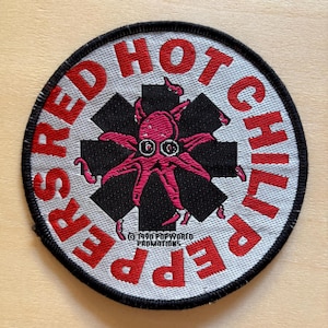 Può includere: Una toppa ricamata circolare con sfondo bianco e bordo nero. Il testo RED HOT CHILI PEPPERS è ricamato in rosso lungo il bordo. Al centro si trova una forma a stella nera con un grafico di un polpo rosa e la scritta 1990.