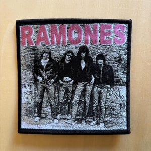 Pode incluir: Um emblema bordado quadrado apresentando uma imagem fotográfica em preto e branco da banda Ramones em pé em uma fila. A palavra RAMONES está bordada em letras maiúsculas rosas grandes e em negrito no topo. O emblema tem uma borda preta espessa.