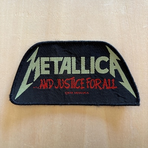 May include: A black fabric patch featuring the Metallica logo in light green embroidery. Below the logo, the text ...AND JUSTICE FOR ALL is embroidered in red. The bottom center includes the small text 1991 METALLICA in red thread.