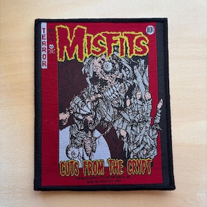May include: A rectangular patch with a black border features a comic book-style design. The word "Misfits" is in yellow, with a red background. The image includes a zombie-like figure and the text "Cuts From The Crypt".