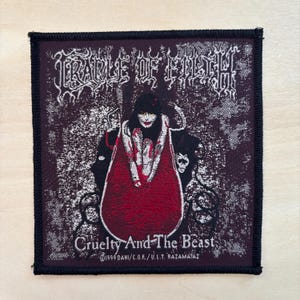 May include: Square patch with the text "CRADLE OF FILTH" in white gothic lettering above a graphic of a woman in a red cloak. The patch also features the text "Cruelty And The Beast" and copyright information.