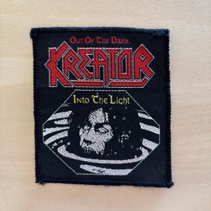 Puede incluir: Un parche rectangular negro bordado que presenta el texto rojo KREATOR con un borde blanco. Sobre el texto está Out Of The Dark en rojo. Debajo hay un gráfico blanco y negro de un rostro emergiendo del agua con el texto Into The Light en amarillo.