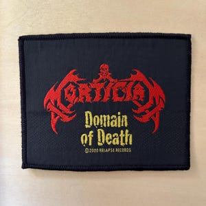 May include: A rectangular black fabric patch with an embroidered red logo reading Mortician, featuring a small skull icon above. Below, the text Domain of Death is embroidered in yellow, followed by the text 2000 Relapse Records.