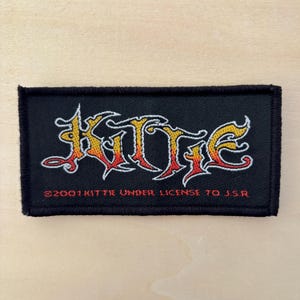 May include: A rectangular black embroidered patch featuring the word Kittie in a stylised, flame-like font with yellow, orange, and red gradients. Below the text, the red embroidered text reads 2001 KITTIE UNDER LICENSE TO J.S.R.