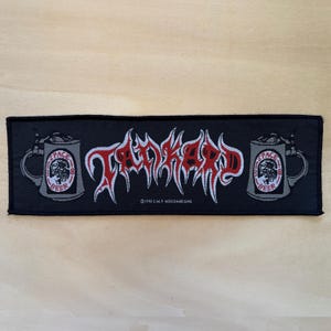 Könnte beinhalten: Ein rechteckiger, schwarz bestickter Aufnäher mit dem rot-weißen Schriftzug TANKARD in stilisierter Schrift. Auf beiden Seiten befindet sich eine graue Bierkrug-Grafik mit einem runden Etikett mit der Aufschrift SPACE BEER. Unten mittig steht 1990 E.M.P MERCHANDISING.