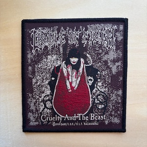 Op de afbeelding: Vierkantige patch met zwarte rand. Het toont de bandnaam "Cradle of Filth" in witte gotische letters. Het ontwerp omvat een donkere achtergrond met een rood-zwarte illustratie van een figuur. De tekst "Cruelty And The Beast" staat eronder.