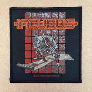Può includere: Toppa quadrata nera con il nome della band "INCUBUS" in lettere arancioni. Sotto, un'illustrazione dettagliata di una figura con abiti rossi e bianchi. La toppa include il testo "©2003 Make Yourself Tour, Inc."
