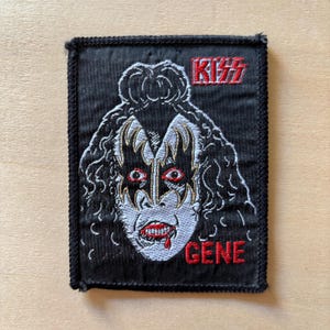 May include: A rectangular black embroidered patch featuring a white and black portrait of a person with theatrical makeup, including black star-shaped eye markings and red lips with a drop of blood. The text KISS and GENE are stitched in red.
